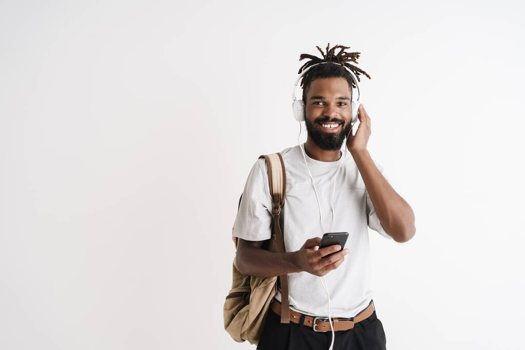 Beyaz arka planda izole edilmiş cep telefonu ve kulaklık kullanan neşeli Afrikalı Amerikalı adamın fotoğrafı. - Fotoğraf, Görsel