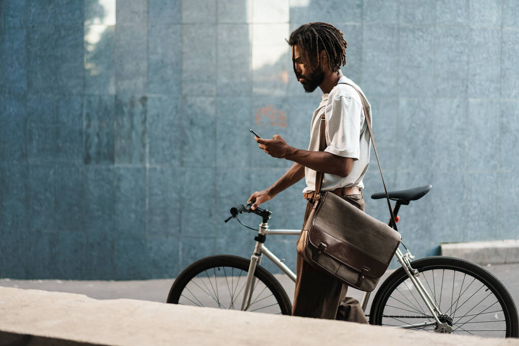 Концентрований афроамериканський хлопець використовує мобільний телефон під час прогулянки з велосипедом на міській вулиці
 - Фото, зображення
