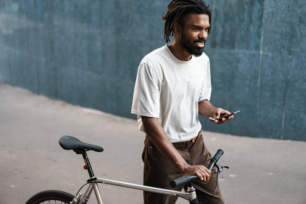 Χαμογελώντας Αφροαμερικανός χρησιμοποιώντας κινητό ενώ περπατούσε με ποδήλατο στην οδό Σίτυ. - Φωτογραφία, εικόνα