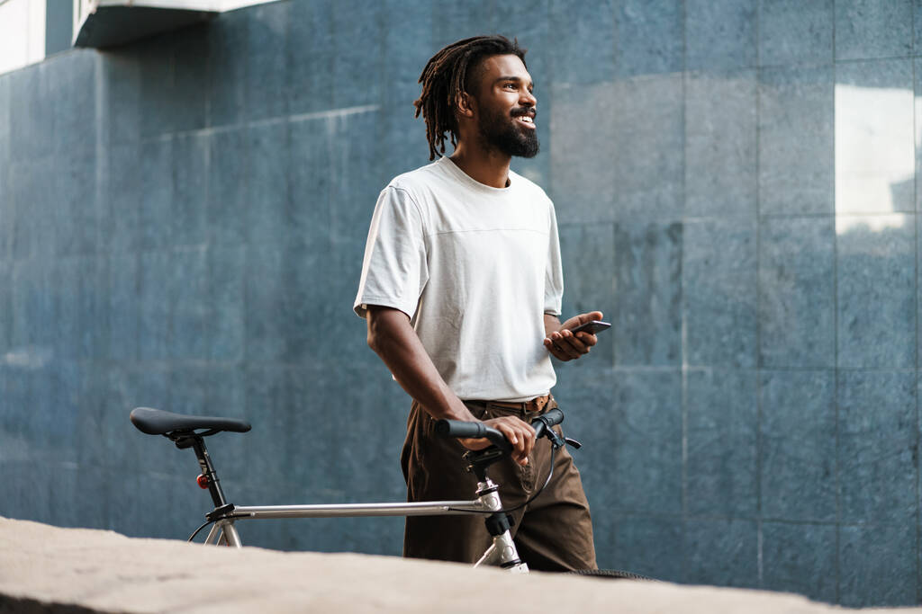 Hymyilevä afrikkalainen amerikkalainen kaveri käyttää kännykkää kävellessään polkupyörän kanssa City Streetillä - Valokuva, kuva