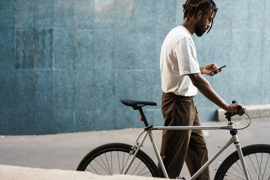 Şehir caddesinde bisikletle yürürken cep telefonu kullanan konsantre Afrikalı Amerikalı adam. - Fotoğraf, Görsel