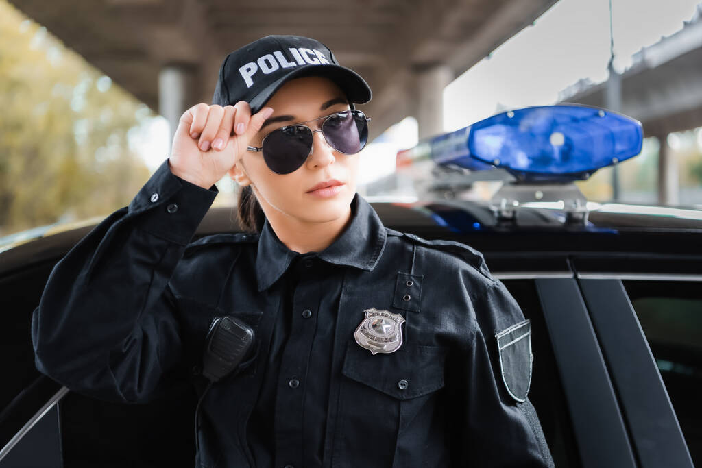 Joven mujer policía mirando hacia otro lado cerca de coche de policía sobre fondo borroso al aire libre  - Foto, Imagen