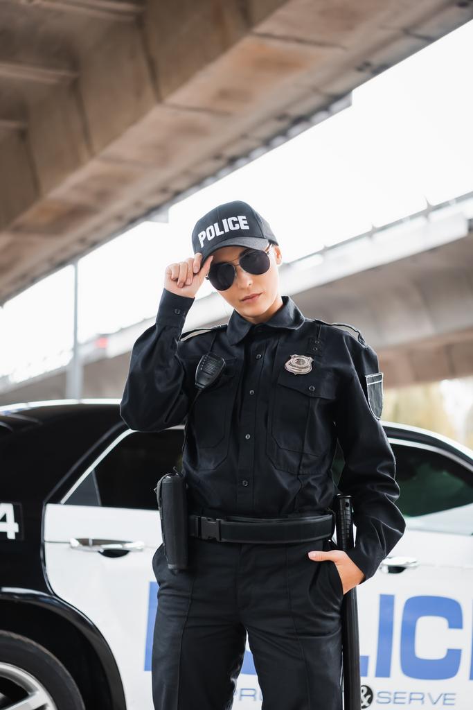 giovane poliziotta con mano in tasca guardando la macchina fotografica vicino pattuglia auto su sfondo sfocato all'aperto - Foto, immagini