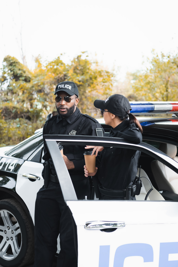 policiais multiculturais com copos de papel falando enquanto se inclina no carro de patrulha no fundo borrado ao ar livre - Foto, Imagem