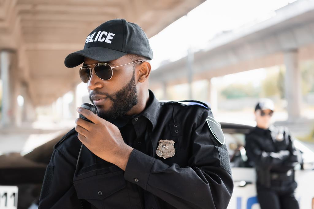 Afrikkalainen amerikkalainen poliisi puhuu radiossa lähellä poliisinaista hämärtyneellä taustalla ulkona - Valokuva, kuva