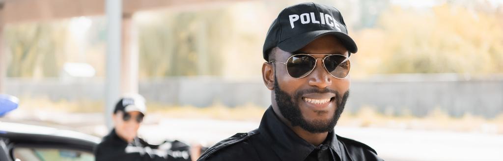 portré boldog afro-amerikai rendőr néz kamera homályos kolléga a háttér szabadban, banner - Fotó, kép