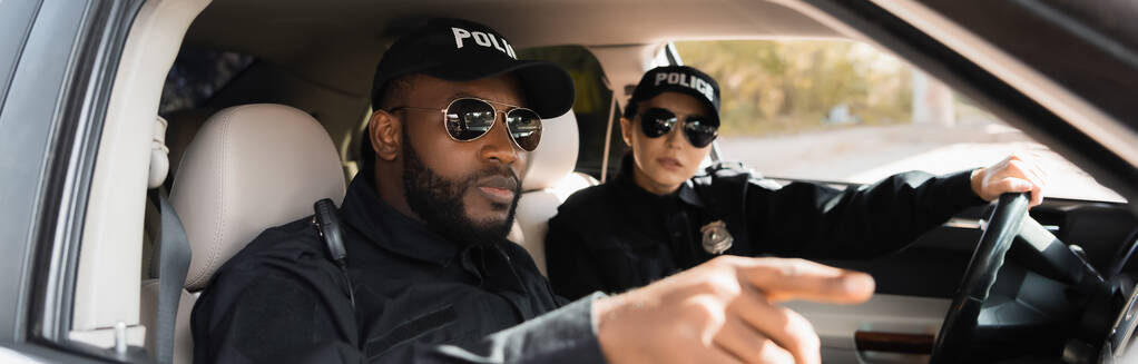 policier afro-américain pointant du doigt près d'un collègue dans une voiture de patrouille sur fond flou, bannière - Photo, image