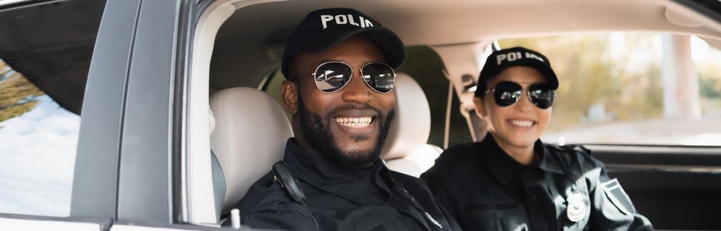 onnellisia monikulttuurisia poliiseja katsomassa kameraa partioautossa hämärtyneellä taustalla, banneri - Valokuva, kuva