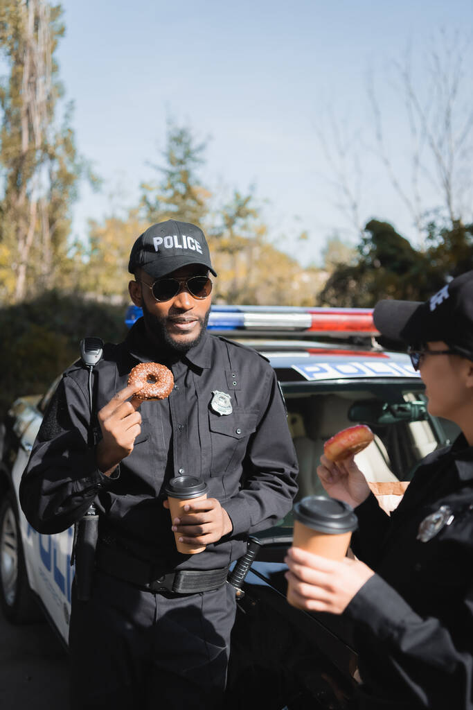 policía afroamericana con rosquilla y taza de papel mirando a colega cerca de patrulla sobre fondo borroso al aire libre - Foto, imagen