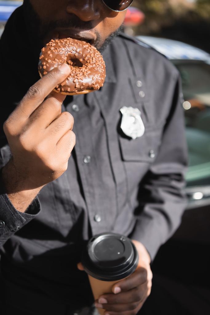 καλλιεργημένη άποψη του Αφροαμερικανού αστυνομικού με χάρτινο κύπελλο τρώγοντας ντόνατ σε θολή φόντο σε εξωτερικούς χώρους - Φωτογραφία, εικόνα