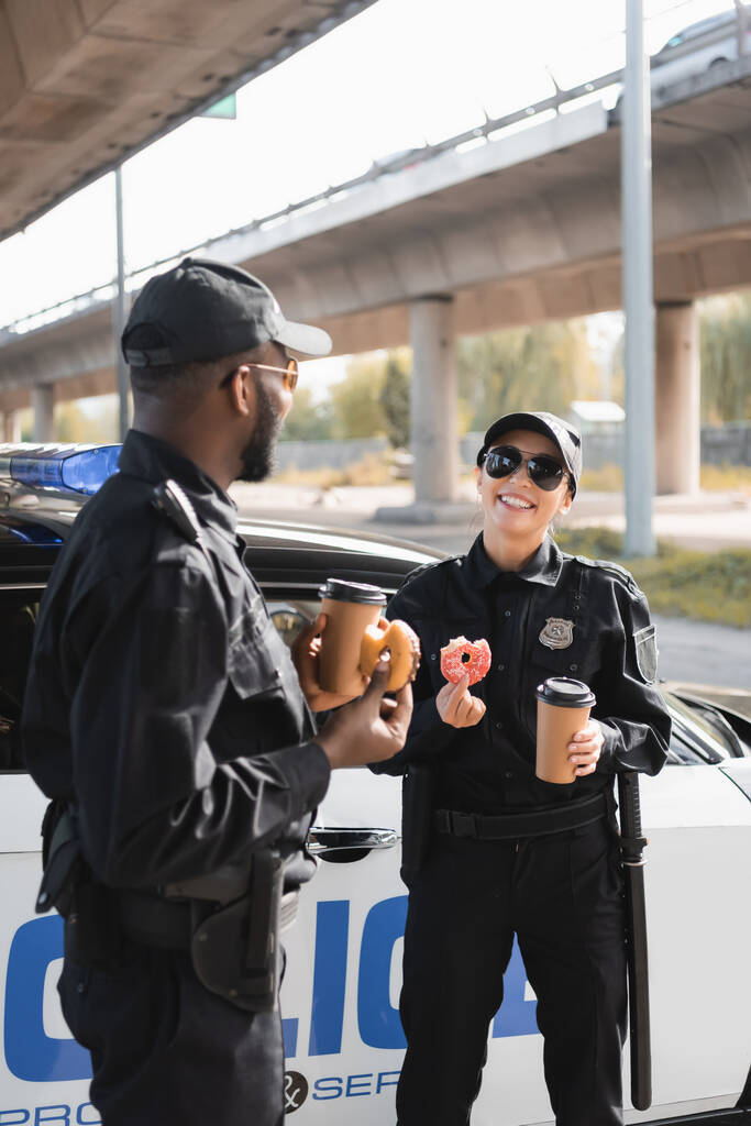 wielokulturowi policjanci z papierowymi kubkami i pączkami rozmawiający w pobliżu radiowozu na rozmytym tle na miejskiej ulicy - Zdjęcie, obraz