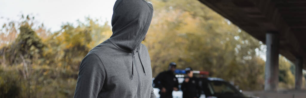 преступник с капюшоном, смотрящий в сторону с размытыми полицией на заднем плане, баннер - Фото, изображение
