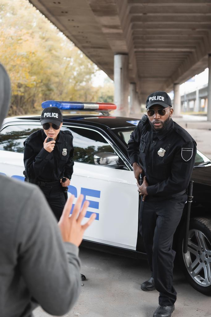 policjantka rozmawiająca przez radio w pobliżu afrykańskiego kolegi wyciągającego broń z zamazanym przestępcą pokazującym dłoń na pierwszym planie - Zdjęcie, obraz