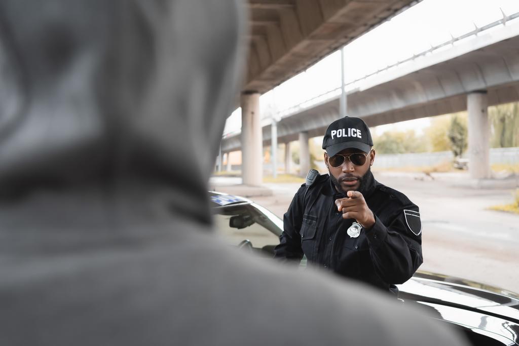 серьезный африканский американский полицейский, указывающий пальцем на размытого преступника в капюшоне на переднем плане на городской улице - Фото, изображение