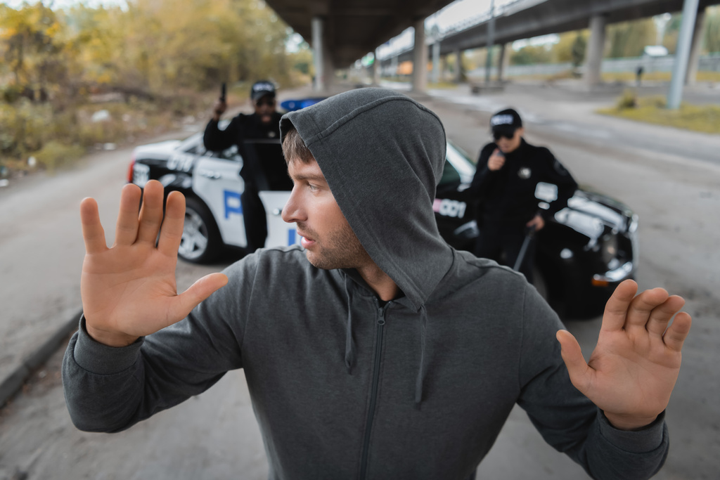 hooded offender with raised hands looking away with blurred multicultural police officers on background on urban street - Photo, Image