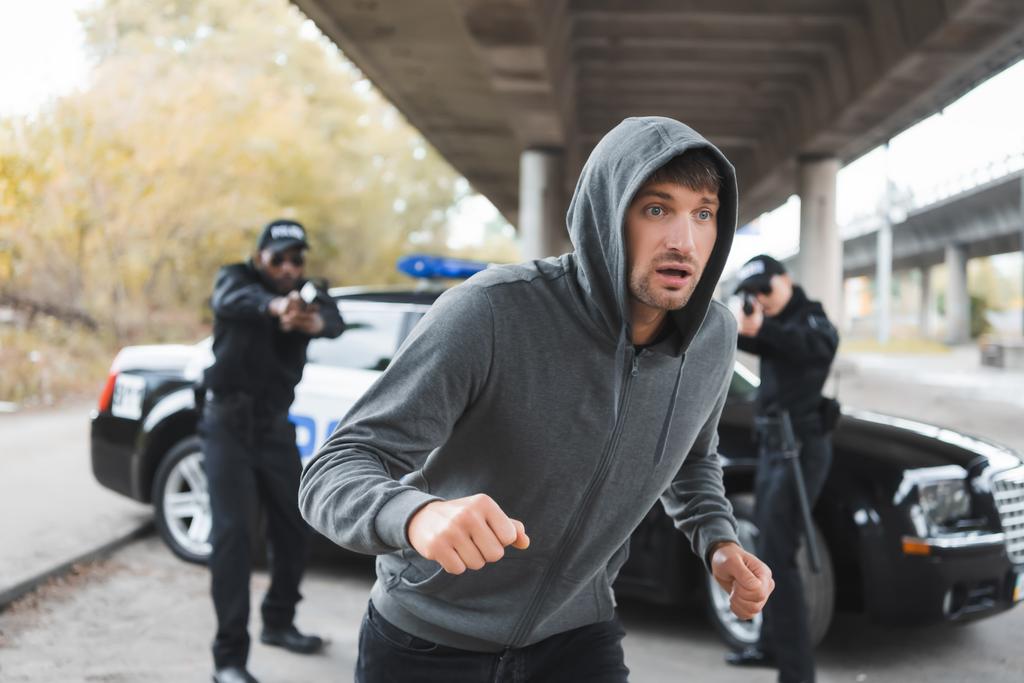 vyděšený pachatel s kapucí utíkání od multikulturní policie míří pistolí na rozmazané pozadí venku - Fotografie, Obrázek