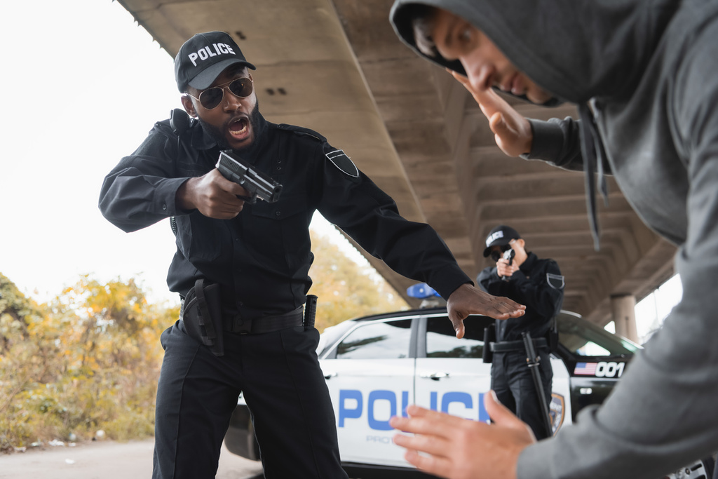 rozzlobený africký americký policista mířící pistolí na rozmazané odevzdané pachatele na popředí v městské ulici - Fotografie, Obrázek