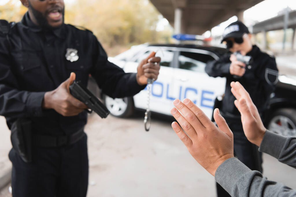 elkövető kezet mutat, miközben multikulturális rendőrök pisztolyt és bilincset tartanak homályos háttéren a szabadban - Fotó, kép