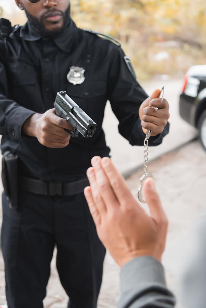 Ausgeschnittene Ansicht eines afrikanisch-amerikanischen Polizisten mit Handschellen, der mit einer Waffe auf den unscharfen Täter im Vordergrund im Freien zielt - Foto, Bild
