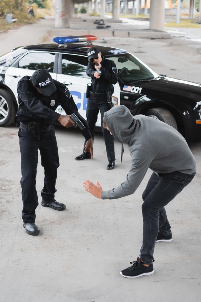 wysoki kąt widzenia wielokulturowych funkcjonariuszy policji z pistoletami wymierzonymi w poddanych przestępców na niewyraźnym tle na zewnątrz - Zdjęcie, obraz