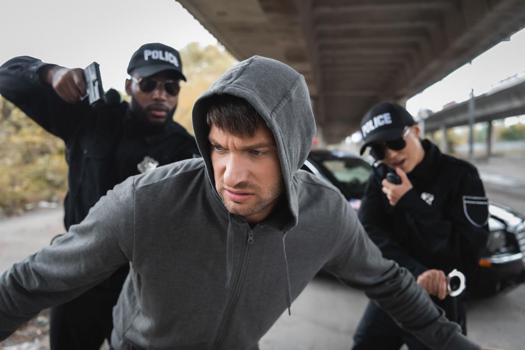dissatisfied hooded offender running from multicultural police officers on blurred background on urban street - Photo, Image