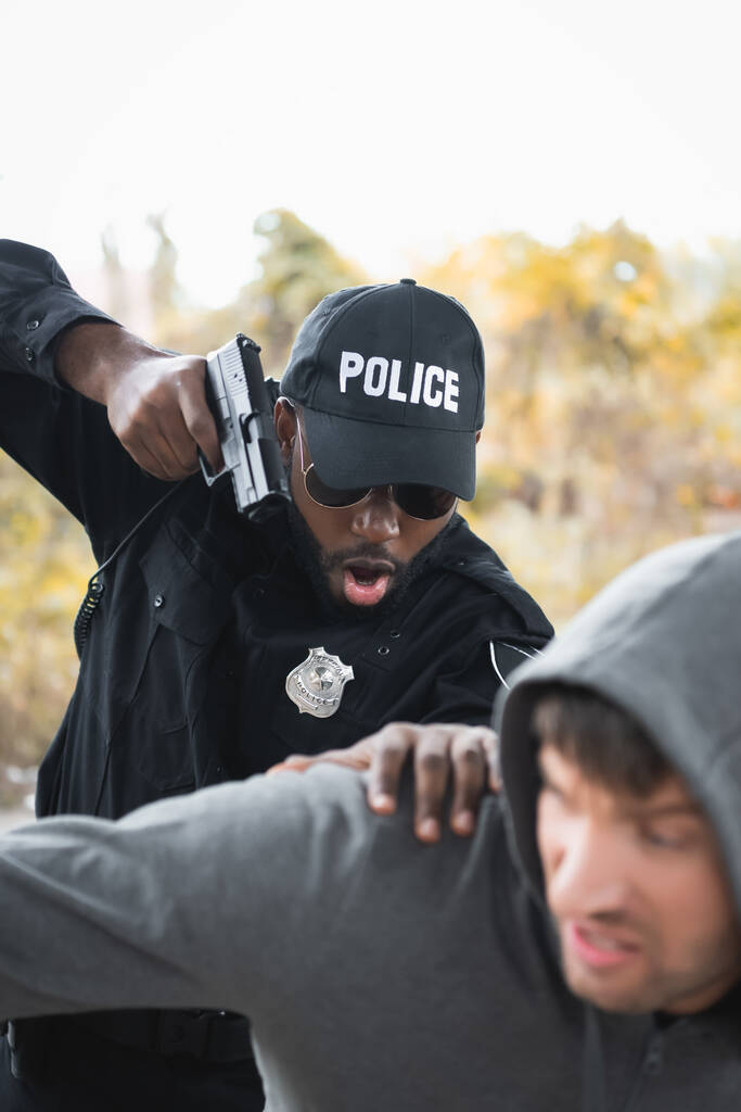 agresivo policía afroamericano apuntando con pistola mientras arresta a delincuente encapuchado en primer plano borroso al aire libre - Foto, imagen