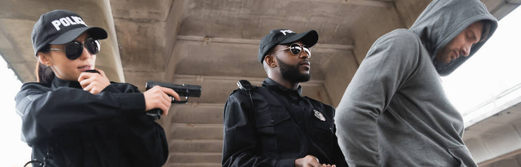 policía afroamericano arrestando a delincuente encapuchado cerca de colega apuntando con pistola al aire libre, pancarta - Foto, imagen