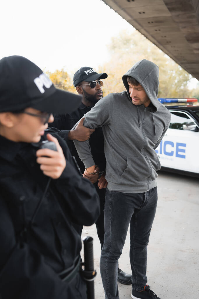 Африканский американский полицейский надевает наручники на преступника в капюшоне с размытым коллегой на переднем плане - Фото, изображение