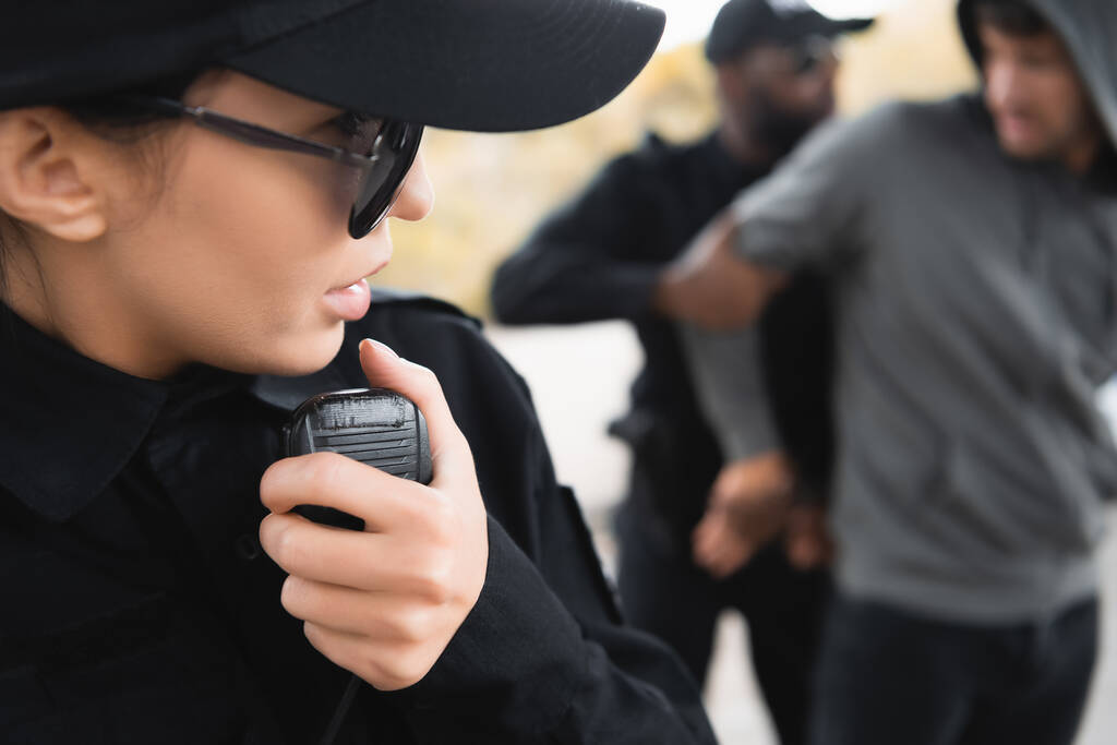vista de cerca de la mujer policía hablando por radio durante colega afroamericano arrestando a delincuente en un fondo borroso - Foto, Imagen