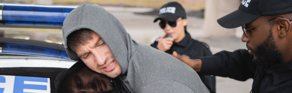 policía afroamericano arrestando a delincuente encapuchado cerca de patrulla con compañero borroso en el fondo al aire libre, pancarta - Foto, imagen