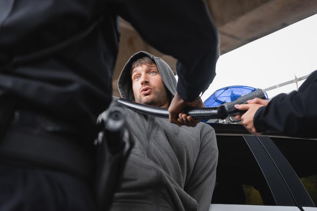 Преступник в капюшоне смотрит на африканского американского полицейского с дубинкой рядом с полицейской, нацеленной пистолетом на размытом переднем плане - Фото, изображение