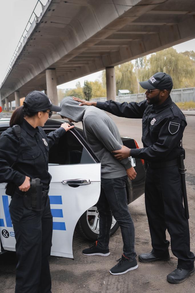 un policier afro-américain arrête un délinquant à capuchon près d'un collègue et d'une voiture de patrouille dans une rue urbaine - Photo, image