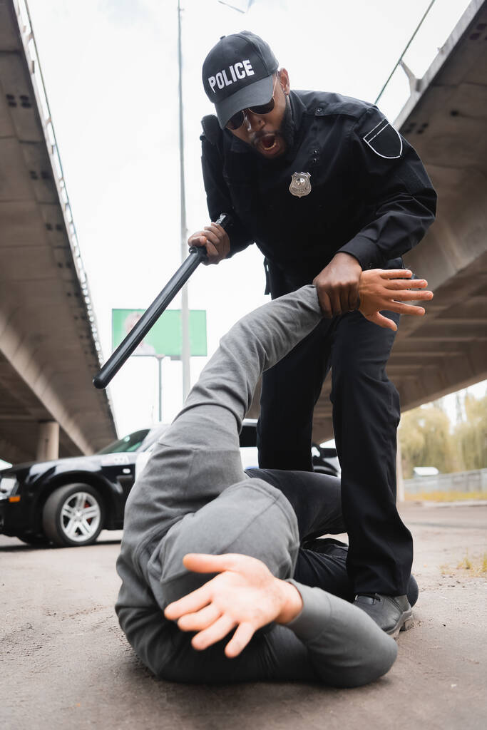 vista de ángulo bajo del oficial de policía agresivo con porra arrestando a delincuente encapuchado acostado en la calle urbana  - Foto, Imagen