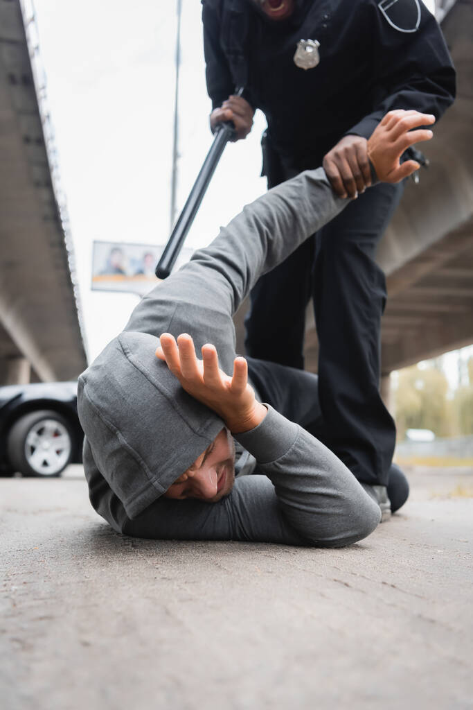 policía afroamericana con porra arrestando a ladrón encapuchado tumbado en la calle sobre fondo borroso - Foto, Imagen