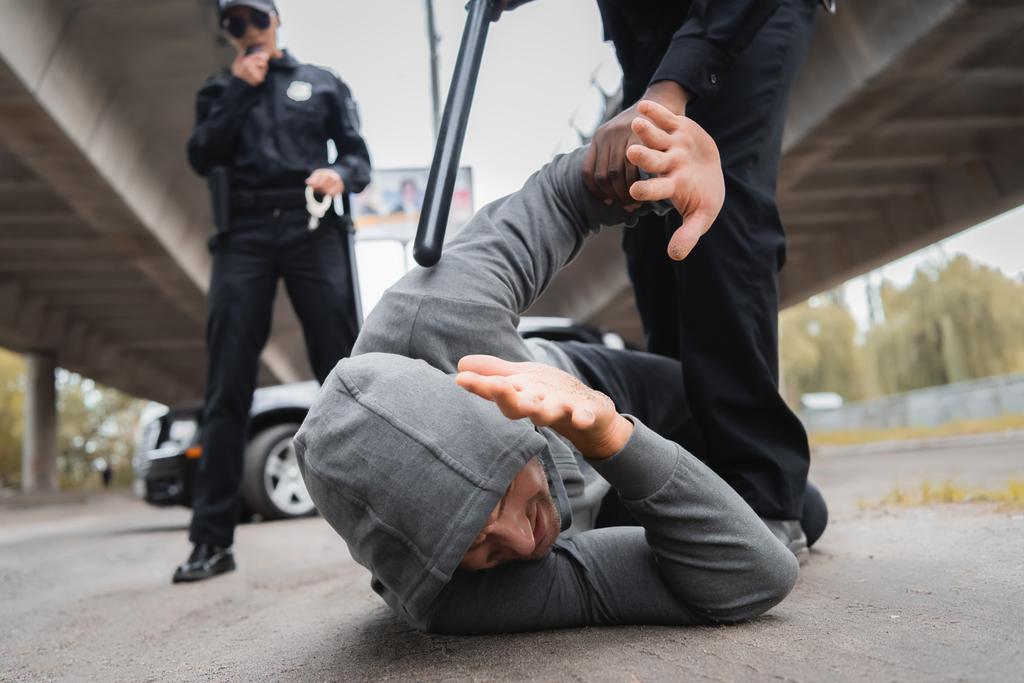 Африканский американский полицейский с дубинкой арестовывает преступника в капюшоне, прикрывающего лицо, пока лежит на улице на размытом фоне - Фото, изображение