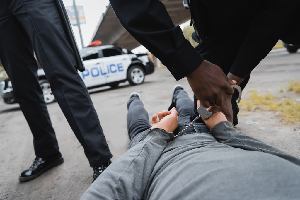 vista recortada del policía afroamericano esposando a delincuente encapuchado acostado en la calle sobre fondo borroso al aire libre - Foto, Imagen