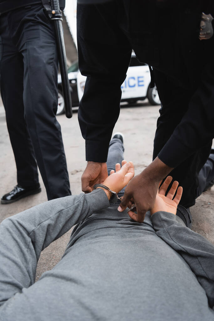 アフリカ系アメリカ人の警察官が同僚の近くの通りに横たわっていた犯人を屋外でぼやけた背景で切り取られたビュー - 写真・画像