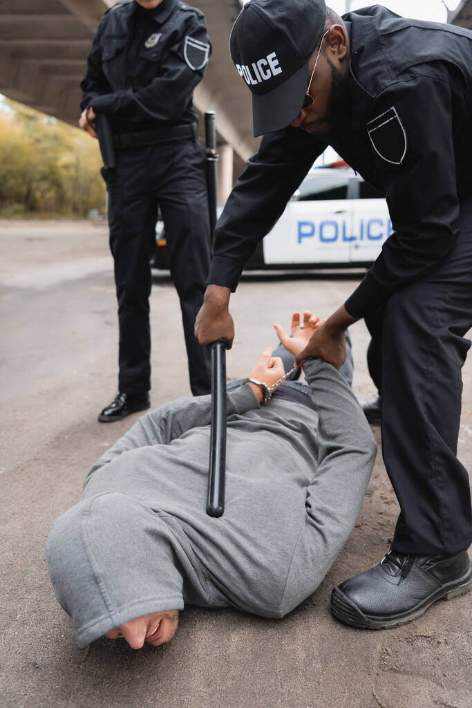 Afrikkalainen amerikkalainen poliisi, jolla on pamppu pidättämässä huppupäistä rikollista makaamassa kadulla hämärtyneellä taustalla. - Valokuva, kuva