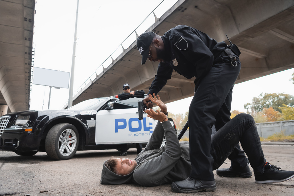 longitud completa del policía afroamericano apuntando con pistola al delincuente asustado que yace en la calle urbana sobre un fondo borroso - Foto, imagen