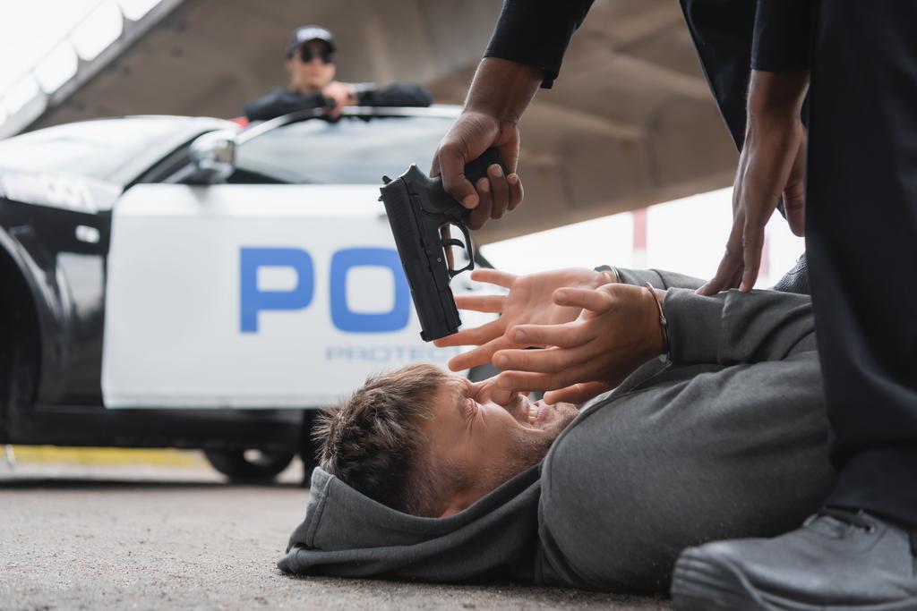 Afrikkalainen amerikkalainen poliisi tähtää pistoolilla pelokkaaseen rikolliseen, joka makaa kadulla hämärtyneellä taustalla. - Valokuva, kuva