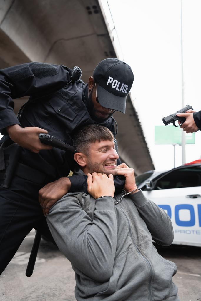 Африканский полицейский-американец душит преступника в наручниках, прицеливаясь пистолетом рядом с коллегой на размытом фоне - Фото, изображение