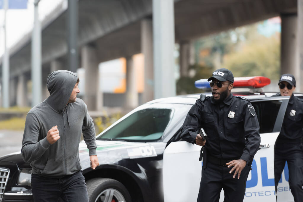 都市部を背景にパトロールカー近くの多文化警察官から逃げるフード犯罪者 ロイヤリティフリー写真 画像素材