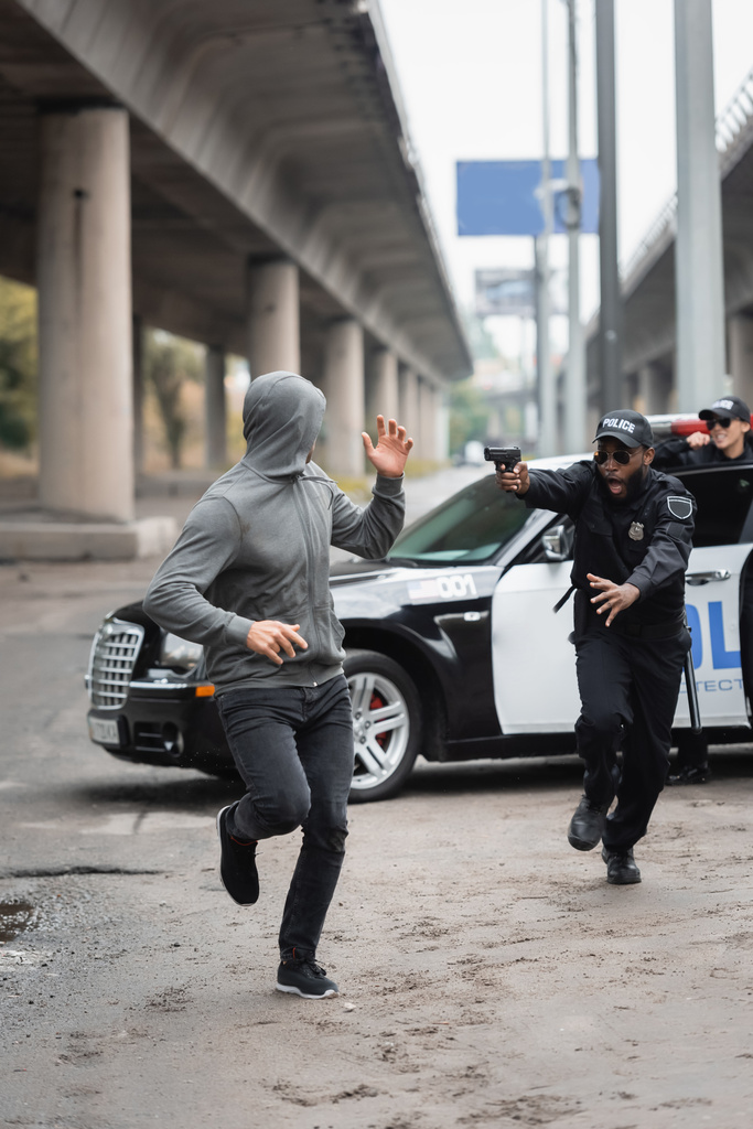 plná délka pachatel s kapucí běží od šokované africký americký policista míří pistolí na rozmazané pozadí na městské ulici - Fotografie, Obrázek
