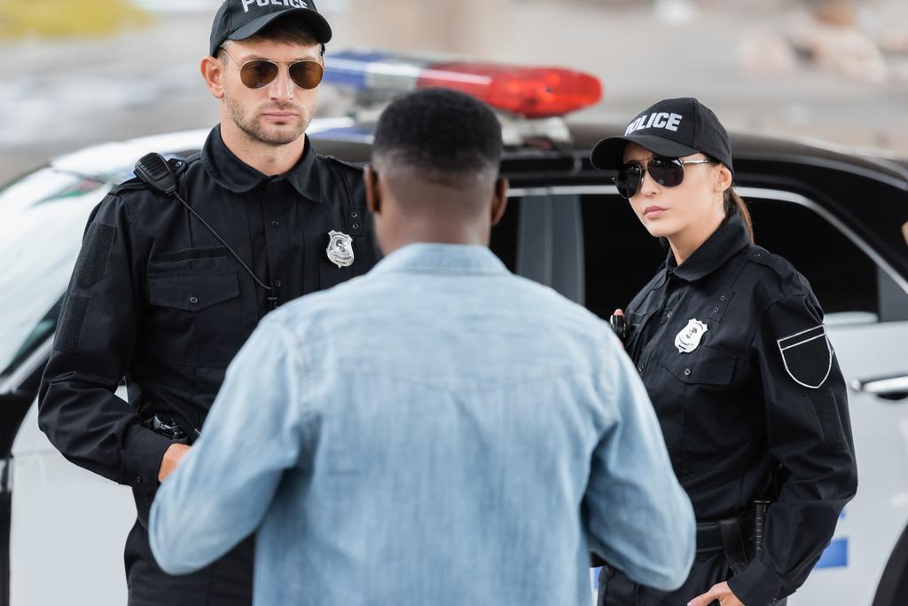 вид ззаду афроамериканської жертви, що стоїть біля серйозних поліцейських на розмитому передньому плані на відкритому повітрі
 - Фото, зображення