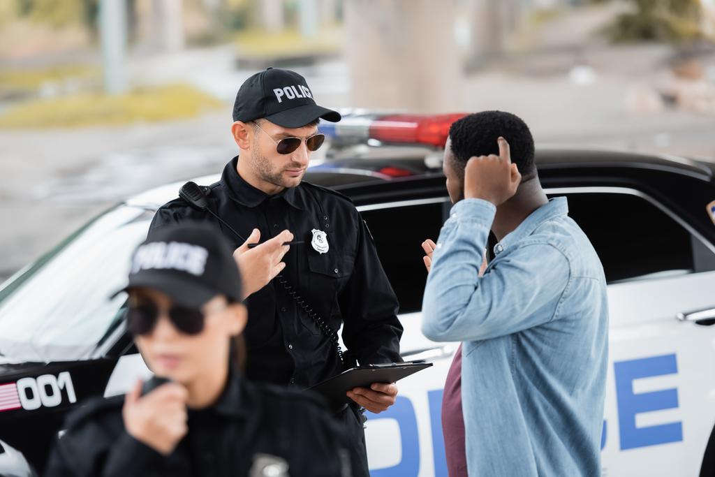 policía hablando con víctima afroamericana cerca de patrulla con colega borroso en primer plano al aire libre - Foto, imagen