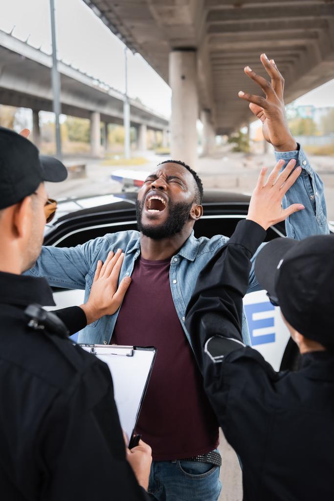 vista posteriore di agenti di polizia calmante disperato uomo africano americano vicino pattuglia auto su sfondo sfocato sulla strada urbana - Foto, immagini