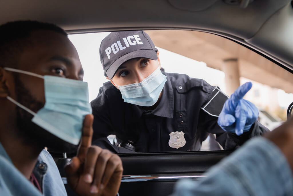 Rendőrnő orvosi maszkban és gumikesztyűben, ujjal mutogatva az afro-amerikai sofőr közelében, aki autóban ül a homályos előtérben.  - Fotó, kép