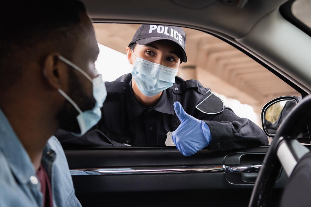 Polizist mit medizinischer Maske und Latex-Handschuh zeigt sich wie afrikanisch-amerikanischer Fahrer im Auto auf verschwommenem Vordergrund  - Foto, Bild