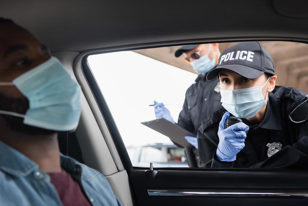 Policia en máscara médica usando walkie talkie cerca de colega con portapapeles y conductor afroamericano en primer plano borroso en coche  - Foto, Imagen
