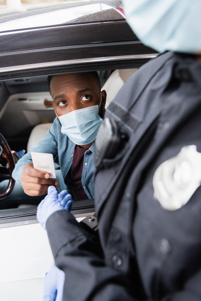 Afrikanischer Fahrer in medizinischer Maske übergibt Führerschein an Polizisten auf unscharfem Vordergrundbild in der Nähe von Auto  - Foto, Bild